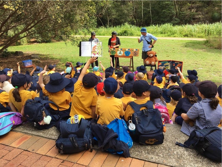 Children sitting on concrete steps looking at some teachers and a big story book